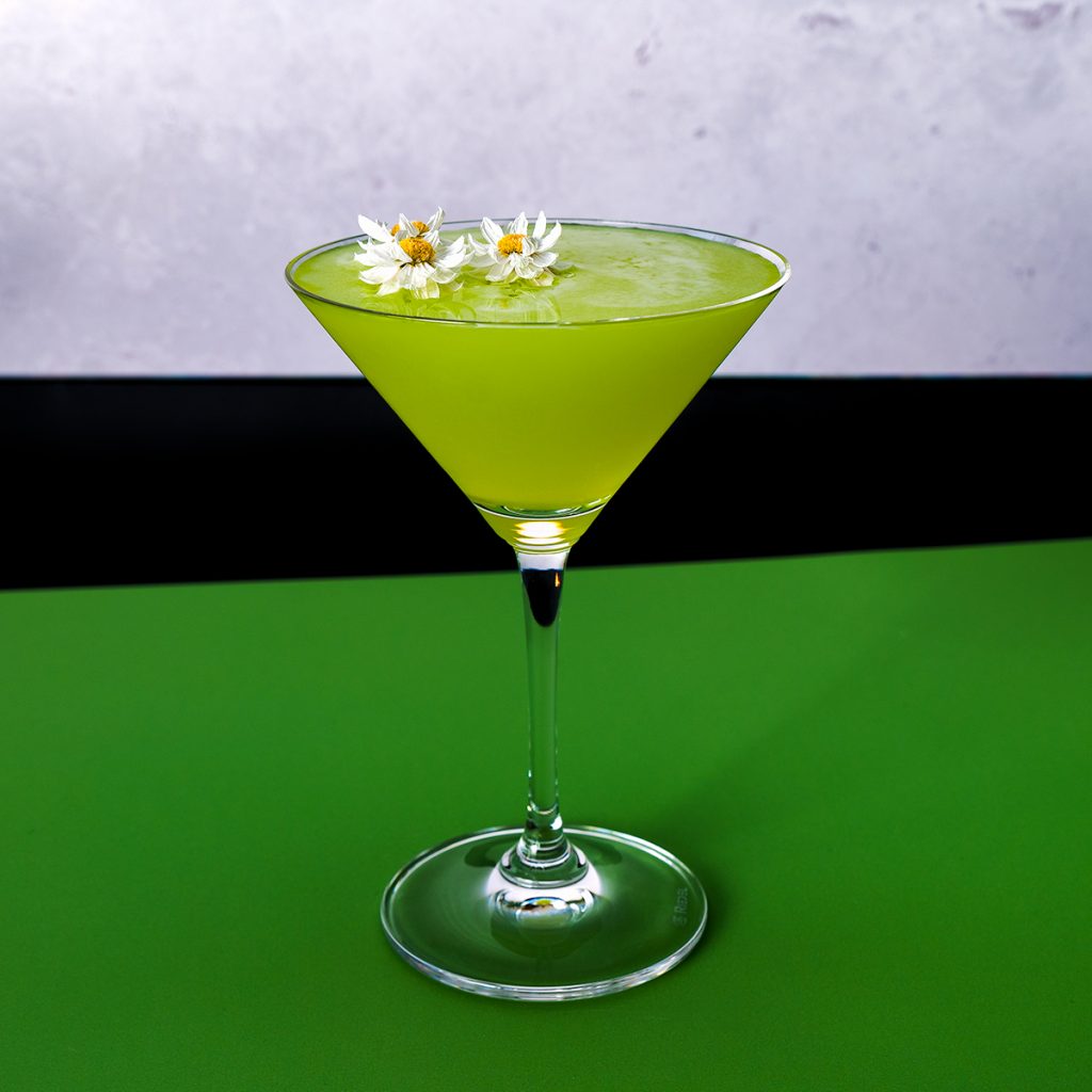 A lime green Spring Stiletto cocktail on a green background, garnished with 3 perfect tiny paper daisies.